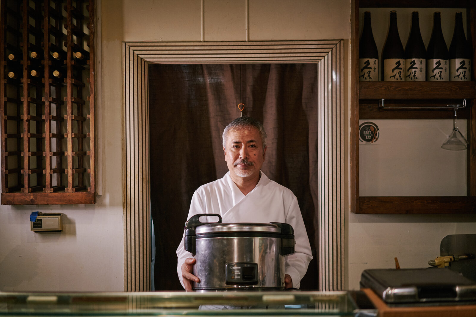A chef stands in the doorway of his sushi restaurant holding a Zojirushi rice cooker, with a chalkboard reading 'We Do Not Carry Any American Style Rolls' beside him